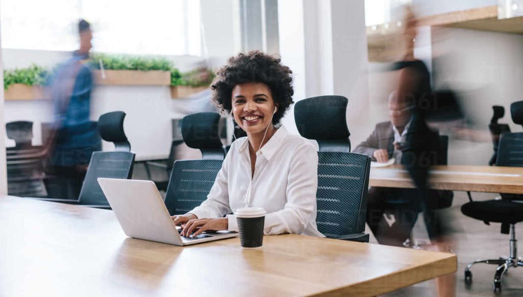Portrait happy young businesswoman working in office. African woman sitting at table with laptop and coffee looking at camera and smiling with people working in background.